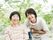 緑の多い屋外で笑顔で過ごしている車いすの高齢女性と付き添いのエプロンを付けた女性スタッフ。