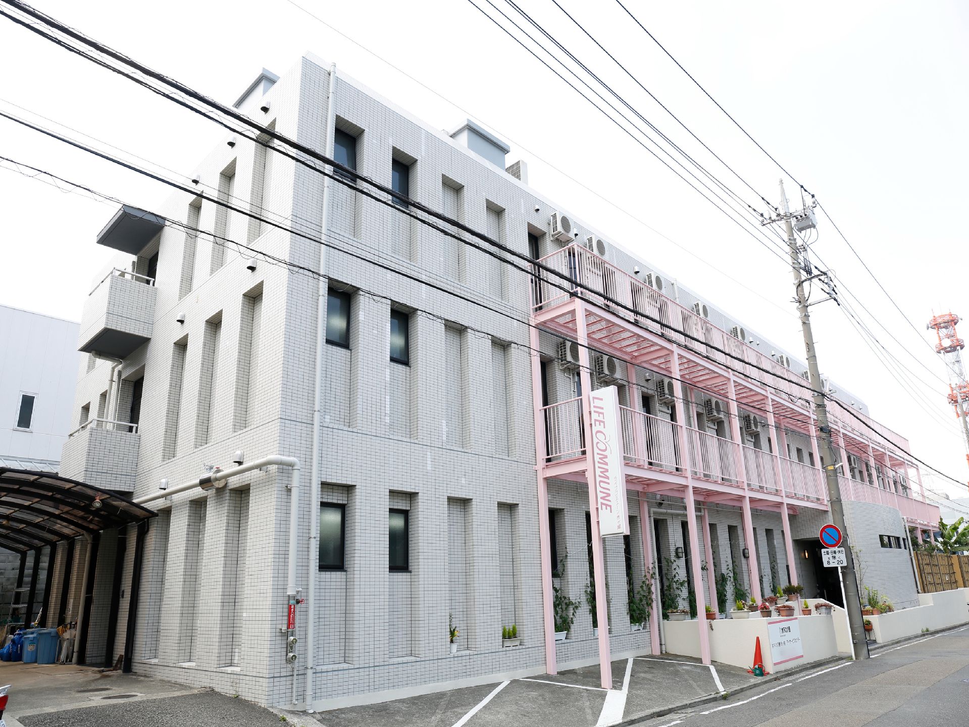 道路に面した立地に3階建てのタイル壁の建物。外階段が設置されている。左側にカーポートがある他、斜めの駐車スペースが2つほど見える。