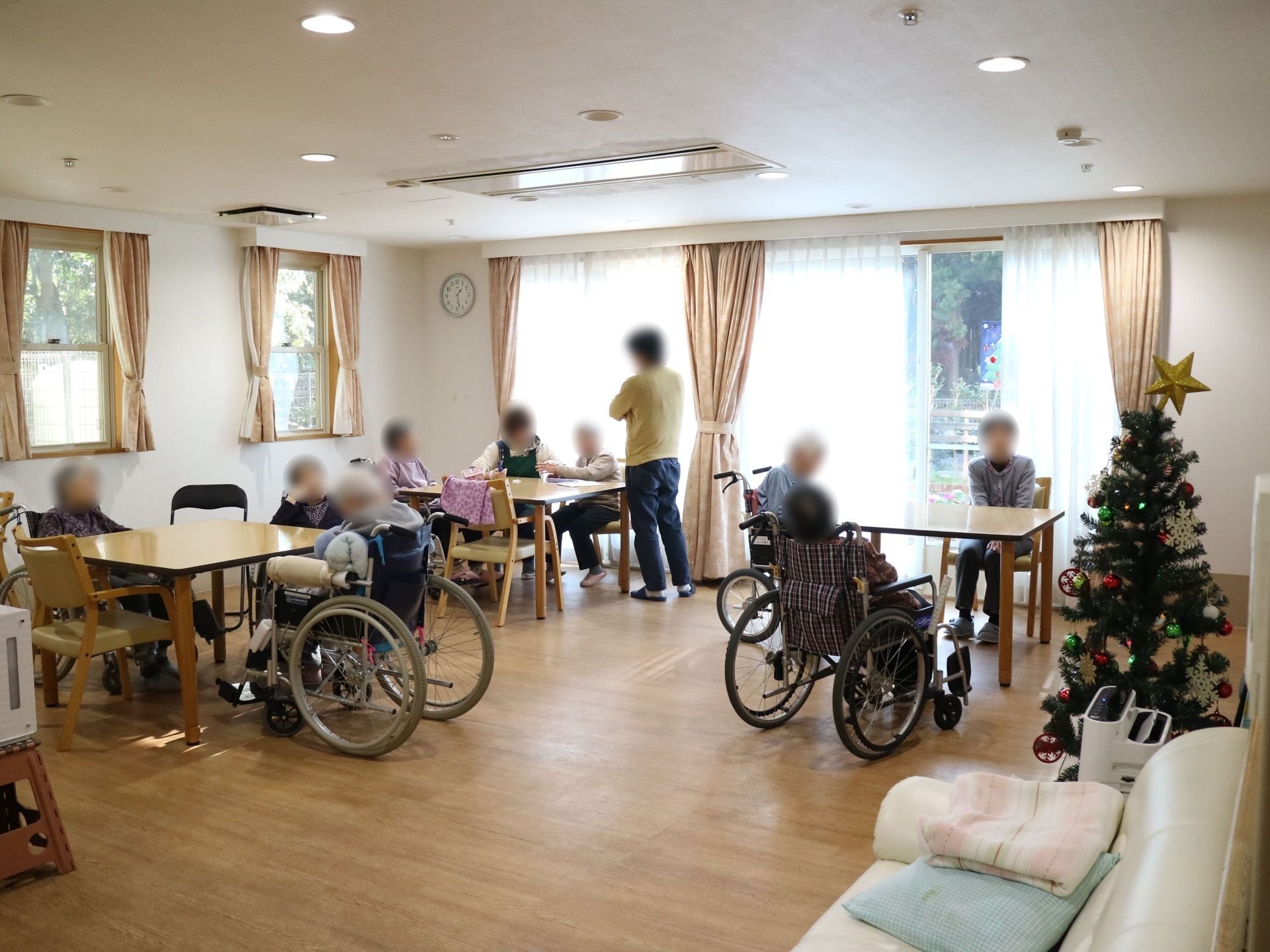 空調設備の整っているリビングダイニングで、天井には複数の照明が設置されている。入居者様やスタッフが席に座って会話をしている。