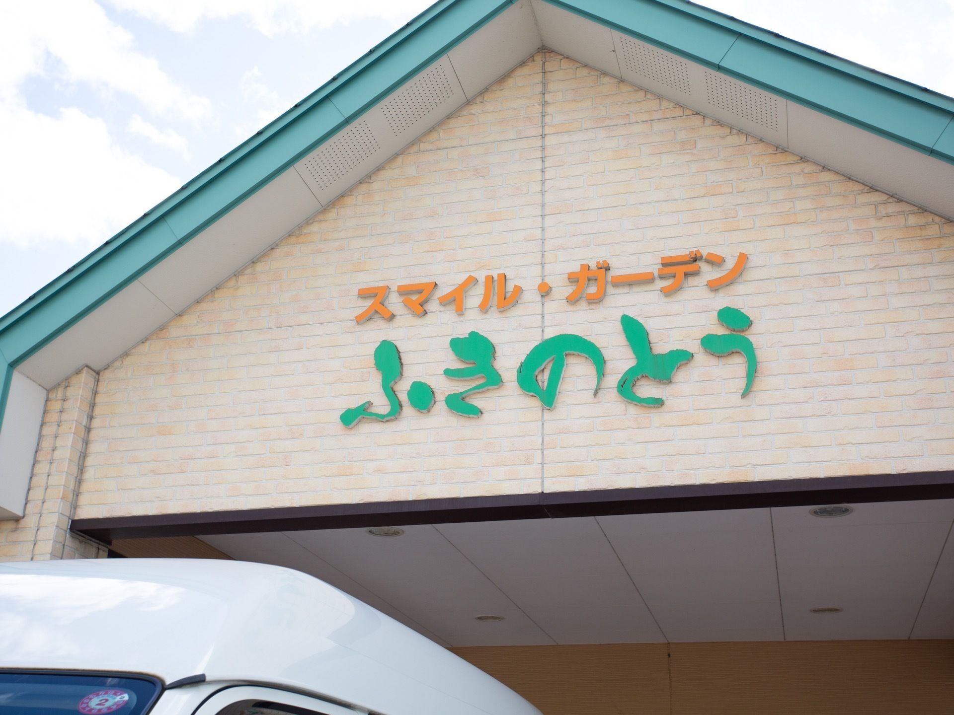 白い壁に水色の三角屋根の付いた施設の玄関がある。黄色とエメラルドグリーンの文字で施設名が大きく書かれている。