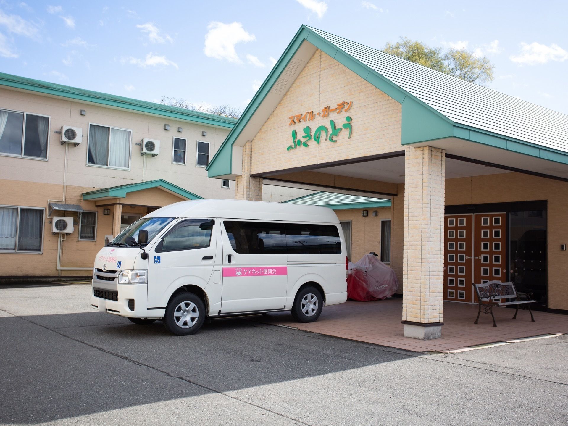 水色の屋根の２階建ての建物がある。玄関の上部分には三角屋根のひさしが付いており、手前には施設の白いワゴン車が停まっている。