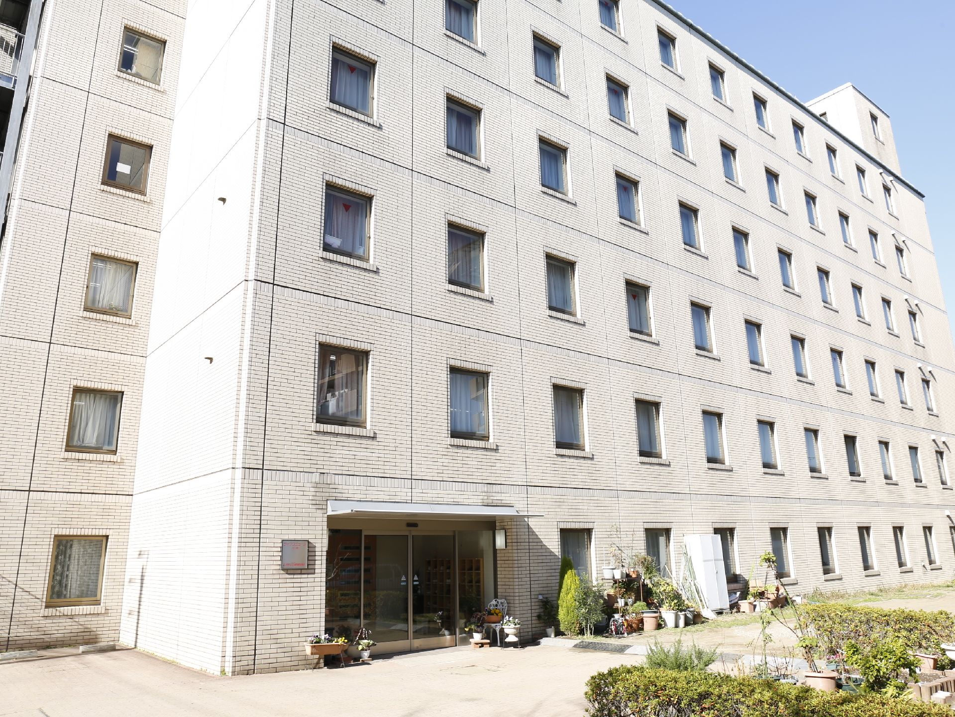 施設は白い外壁で6階建てである。建物の端には非常階段がある。入口付近には植木鉢などが置いてあり、木も植えられている。