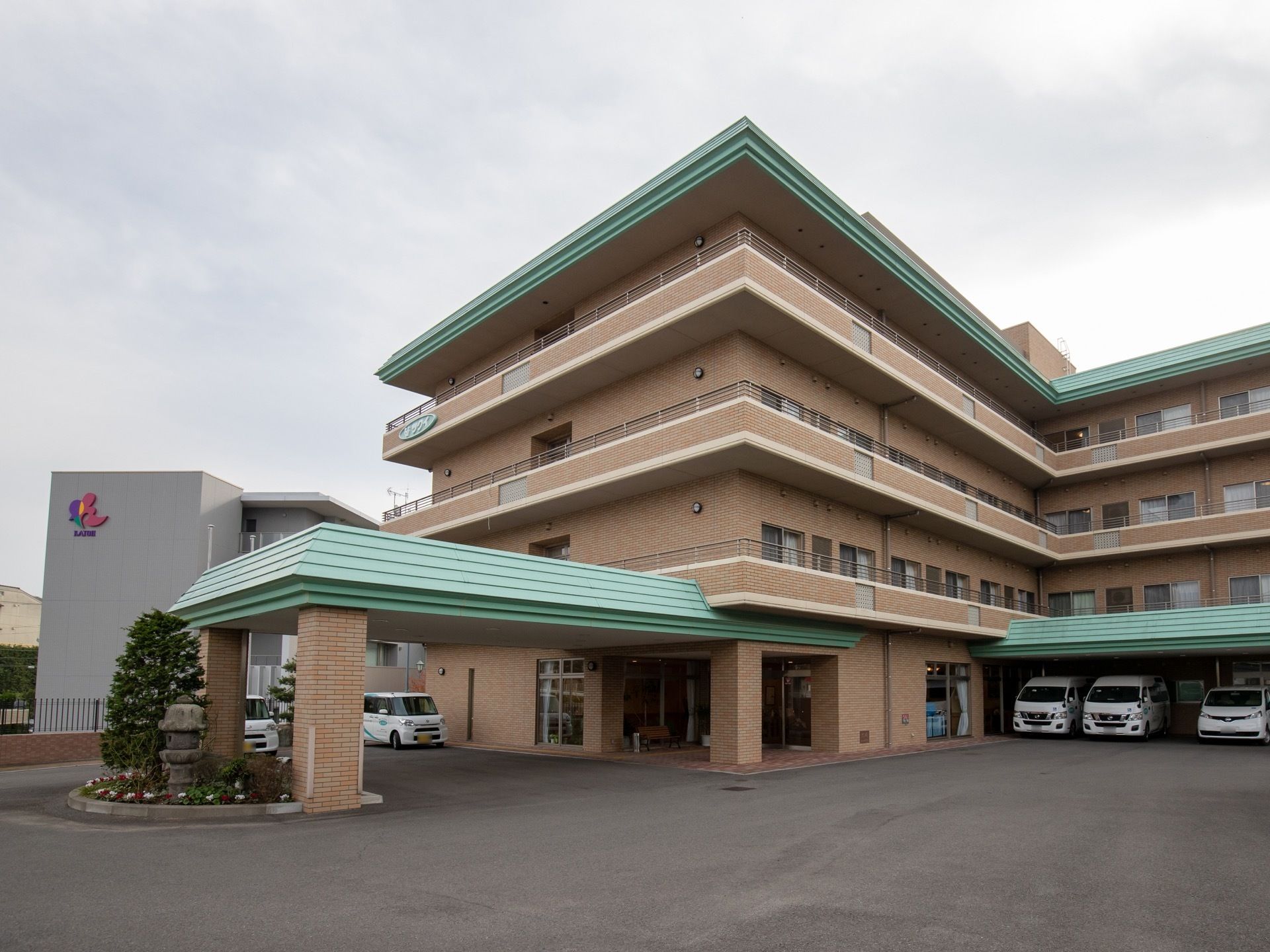 茶色とエメラルドグリーンが使われた落ち着いた外観である。玄関には車寄せがあり、施設の車が置かれたスペースもある。