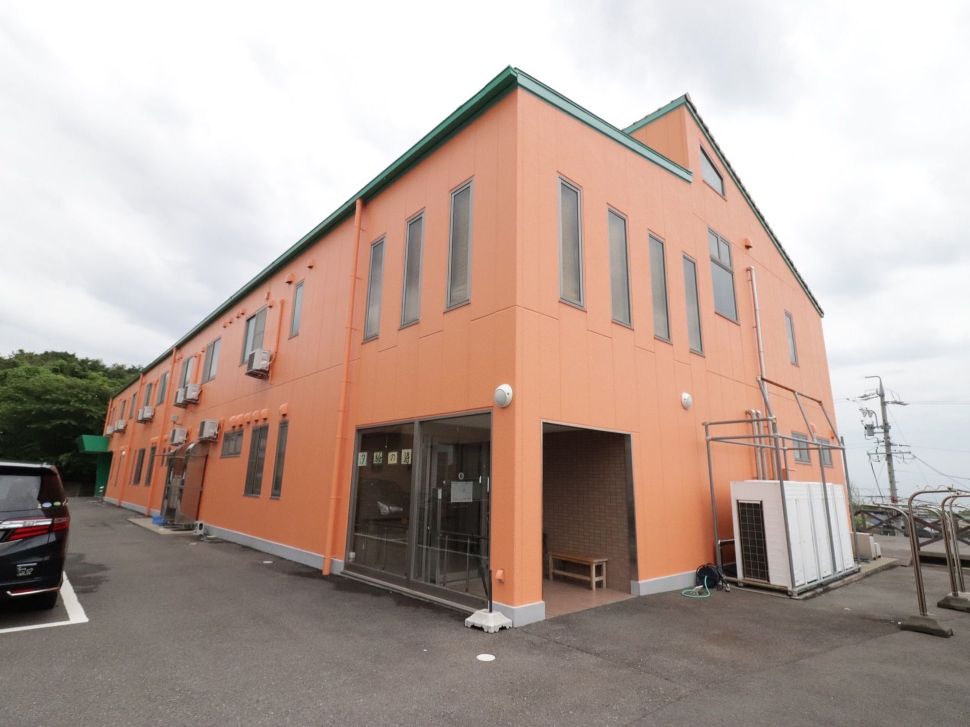 地上２階建ての施設は、ライトオレンジと緑色のツートンカラーである。左横には駐車場があり、黒い車が置いてある。