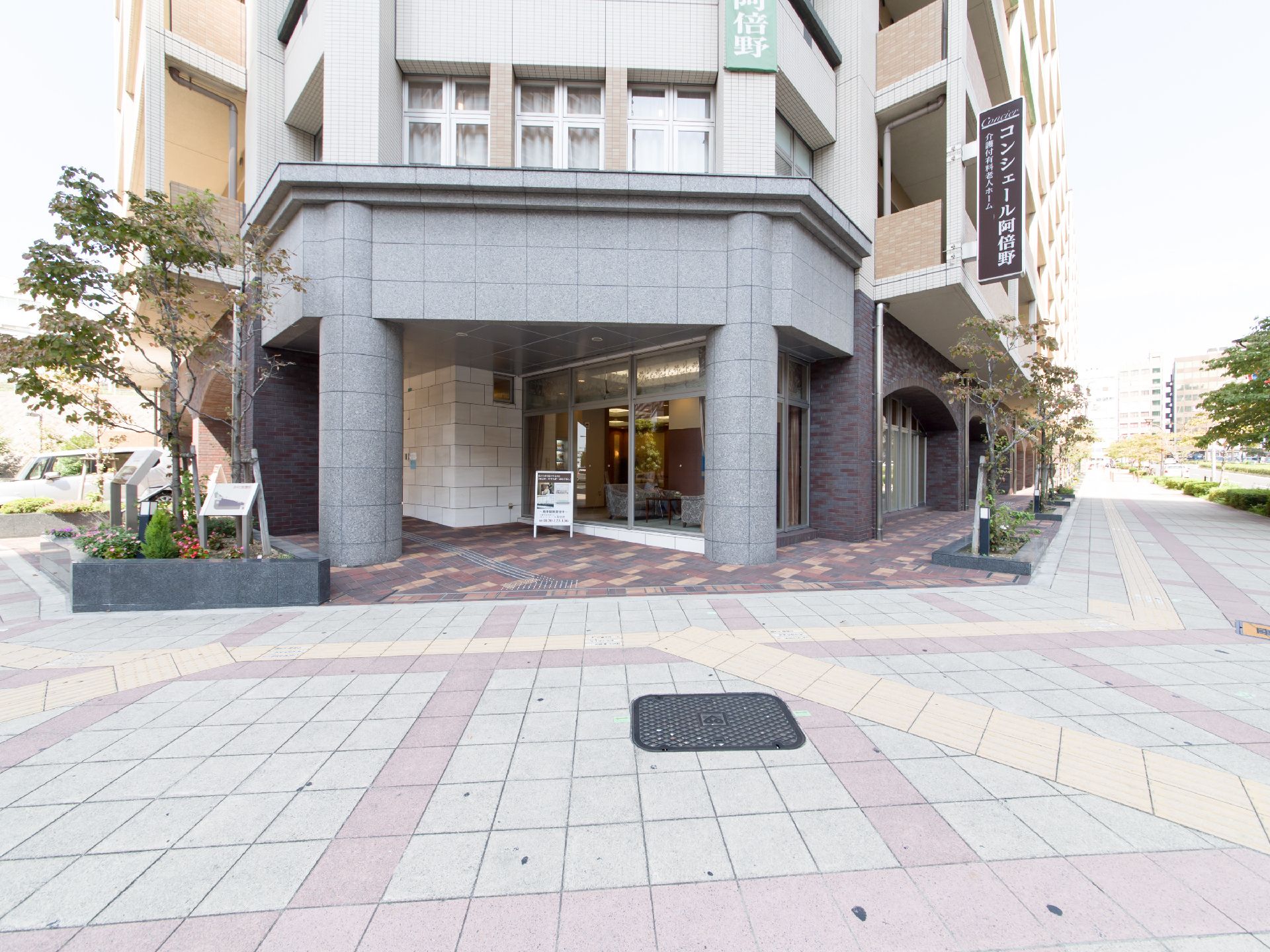 施設1階前には広々とした歩道があり、間隔をあけて木が植えられている。建物2階のところに施設の看板がある。