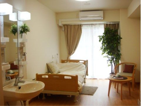 ベッドやテーブルなどが置かれている。窓際に大きな観葉植物があり、手前の洗面台の棚にも小さな植物が飾られている。