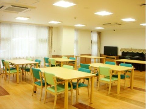 長方形のテーブルが複数あり、それを挟んで緑と黄緑の椅子が１脚ずつ交互に置かれている。奥の壁際にはテレビがある。