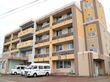 施設の外観はベージュ色を基調としており、１階の一部がレンガ張りの壁で、アクセントになっている。建物の前に車が３台停められている。