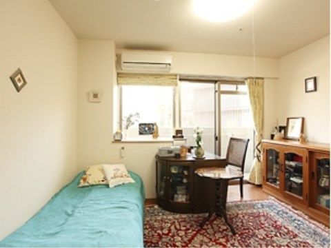 絨毯の敷かれた部屋にベッドが置かれている。窓際と壁際には整理棚などの調度品が並んでいる。入居者様のお気に入りの家具を持ち込むことができる。