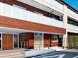 通りに面した３階建ての建物。入口へのアプローチは手すり付きの階段とスロープの両方が設置されているので安心である。