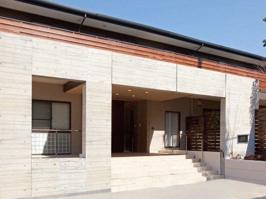 コンクリート造りの頑丈な2階建ての建物。木製フェンスでプライバシーにも配慮。大きな屋根のついた玄関へと続くアプローチも広い。