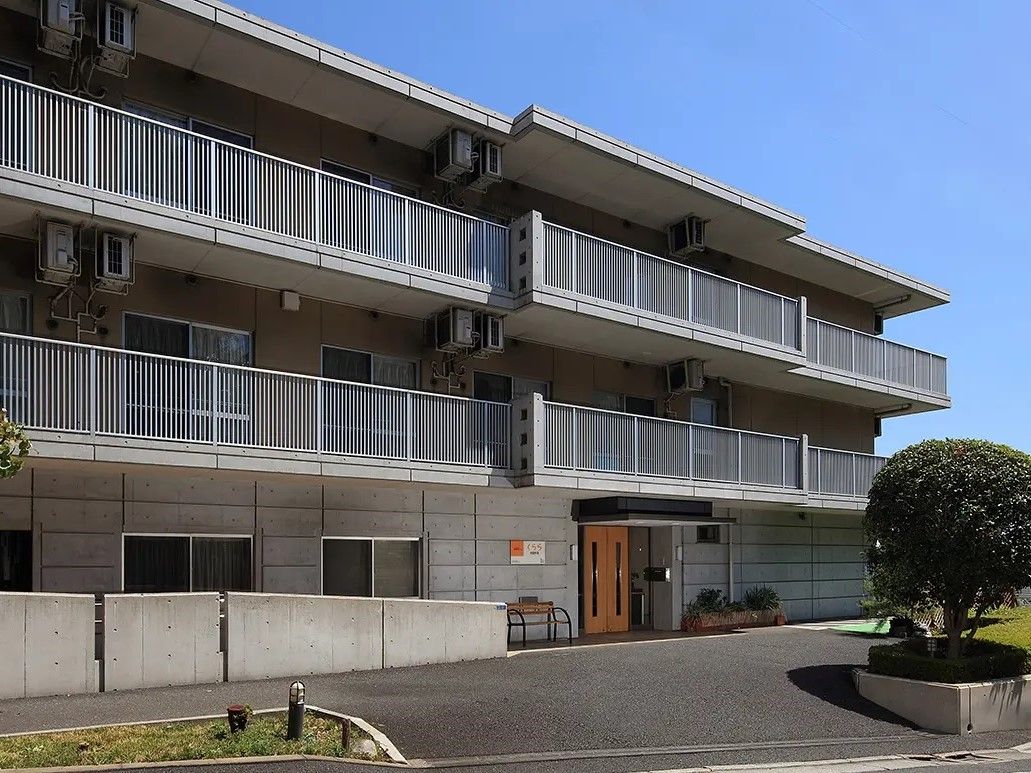 石張りの外壁とコンクリート造りのアプローチがシックなマンション風の建物。敷地への入り口に植え込みがある。