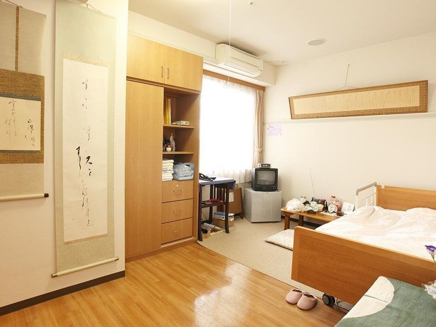 壁に飾られた掛け軸が和の落ち着いた雰囲気を感じさせる居室。ベッドやテレビの置かれたスペースにはマットが敷かれている。