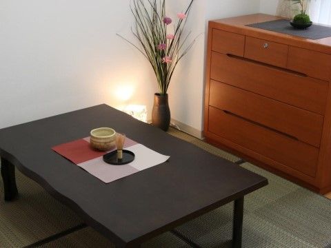 窓際に４段の小型整理ダンスが置いてある。床にはござを敷き褐色の低いテーブルが置かれ、和風に設えてある。壁際に生け花と間接照明もある。