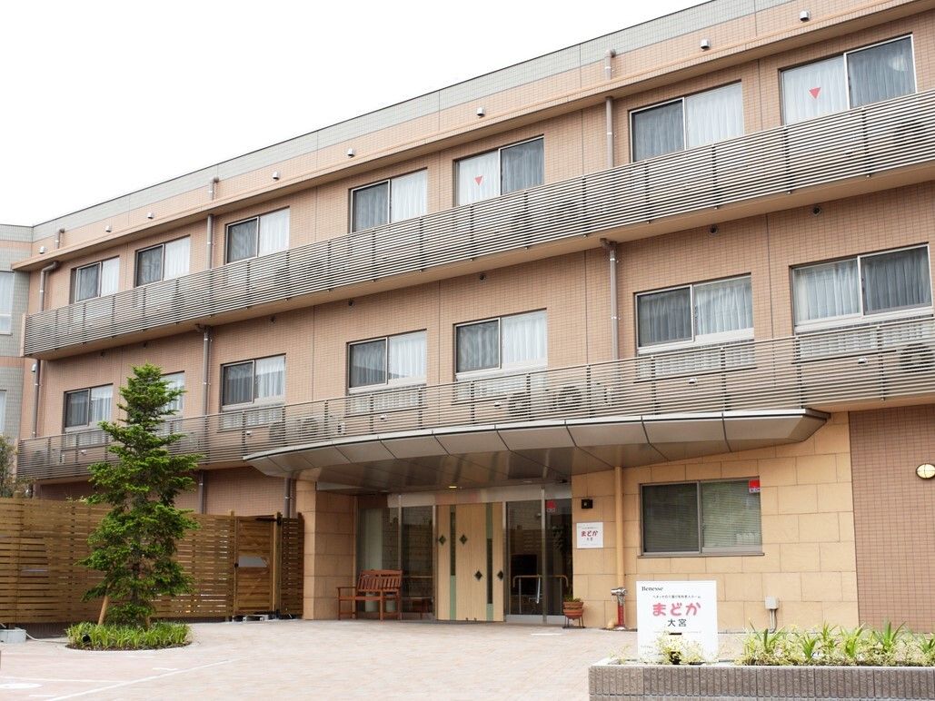 ３階建てでベージュ色の建物は、居室窓の前にはベランダがある。玄関上にはひさしがあり、敷地入口から玄関までは舗装されている。