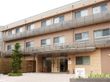 ３階建てでベージュ色の建物は、居室窓の前にはベランダがある。玄関上にはひさしがあり、敷地入口から玄関までは舗装されている。