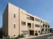 明るいブラウン色の大きな建物。三階建てで、入口には屋根が付いている。手前は駐車スペースになっている。