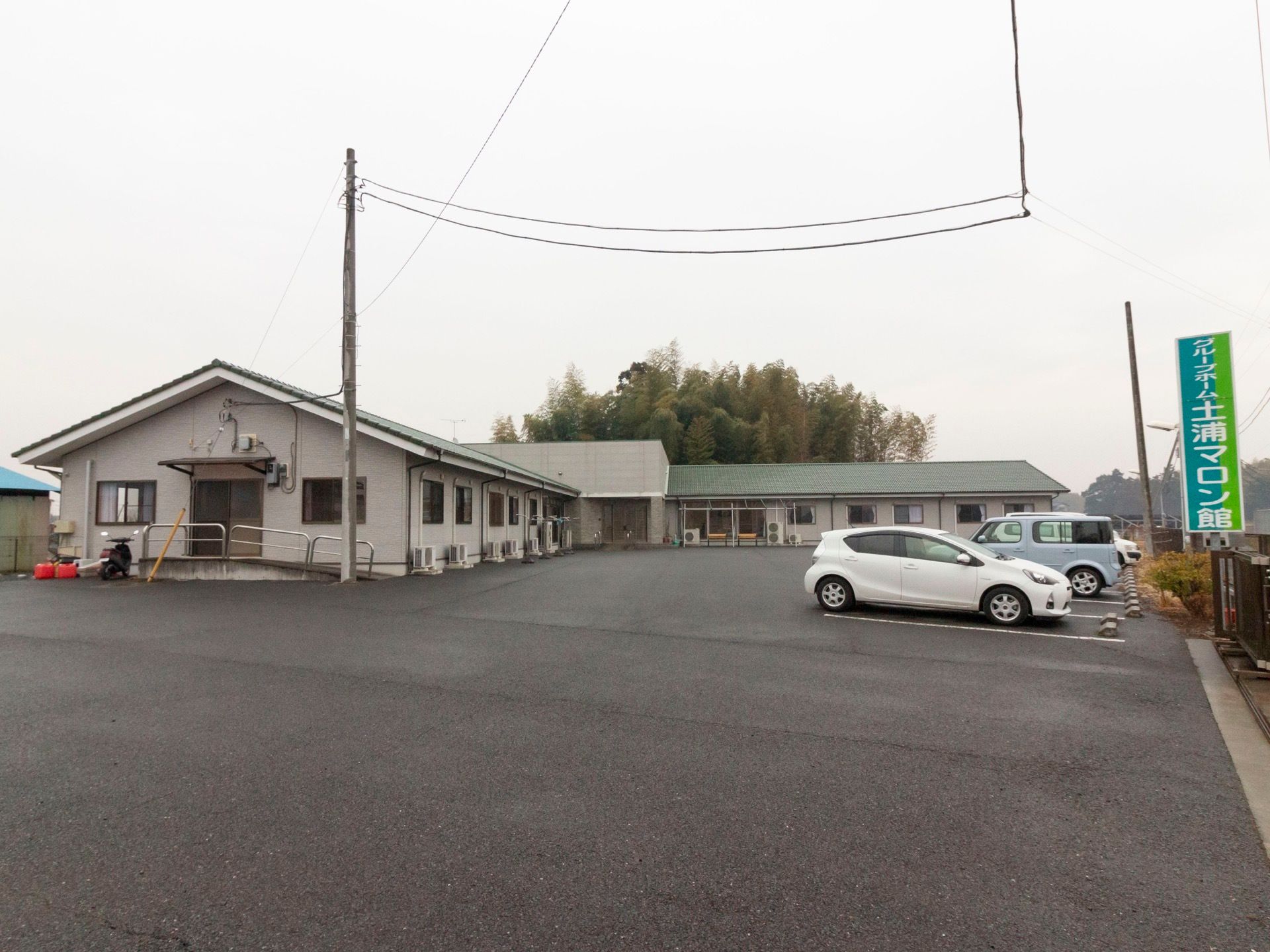 施設は平屋建てである。電柱のすぐそばには扉があり、緩やかなスロープと手すりが設置されている。敷地内にはバイクや車が停められている。