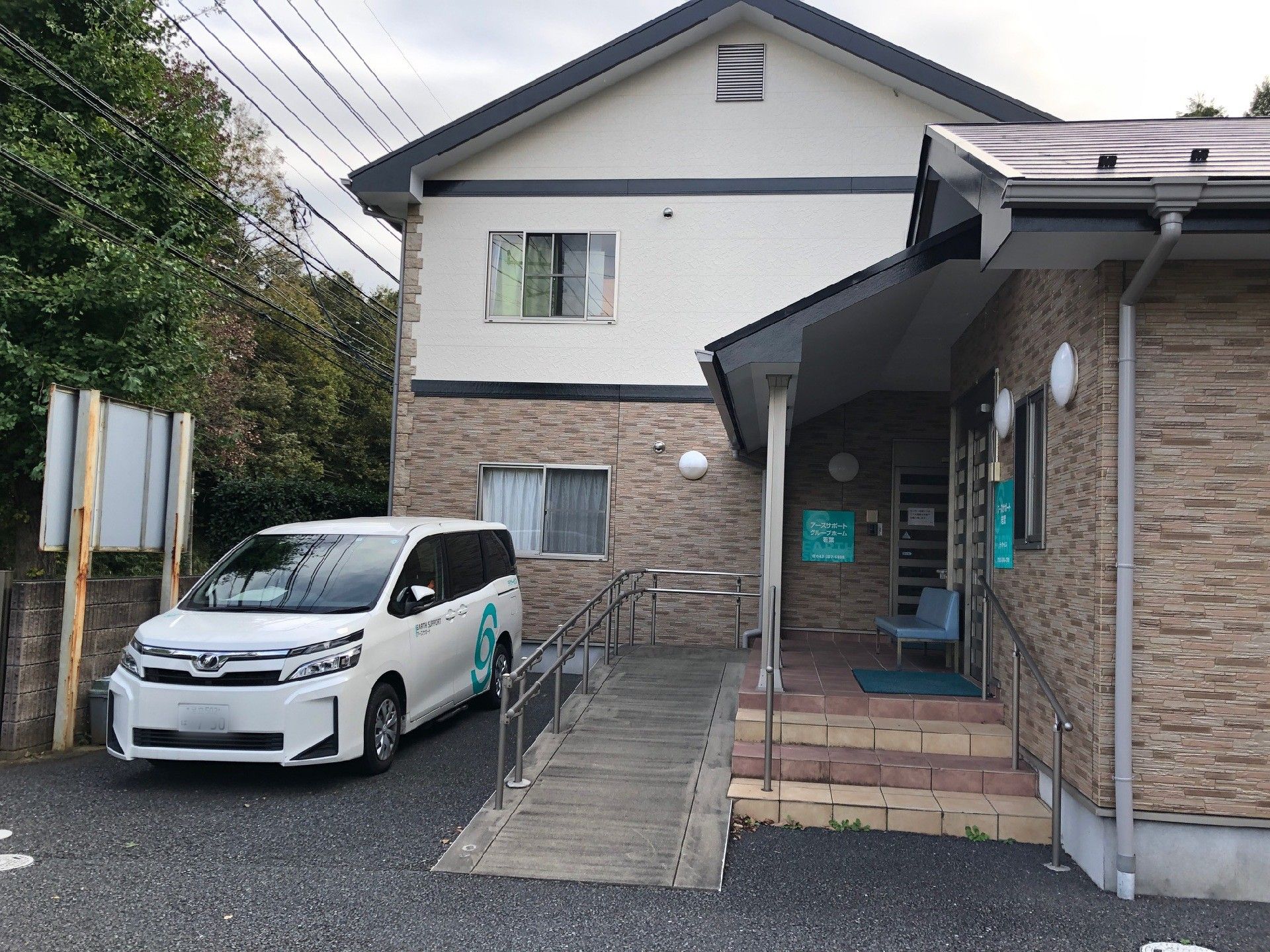 居住部分とデイサービスは別の建物で、エントランスの通路でつながる形になっている。タイル調のおしゃれな外観で、敷地内に送迎車が停められている。