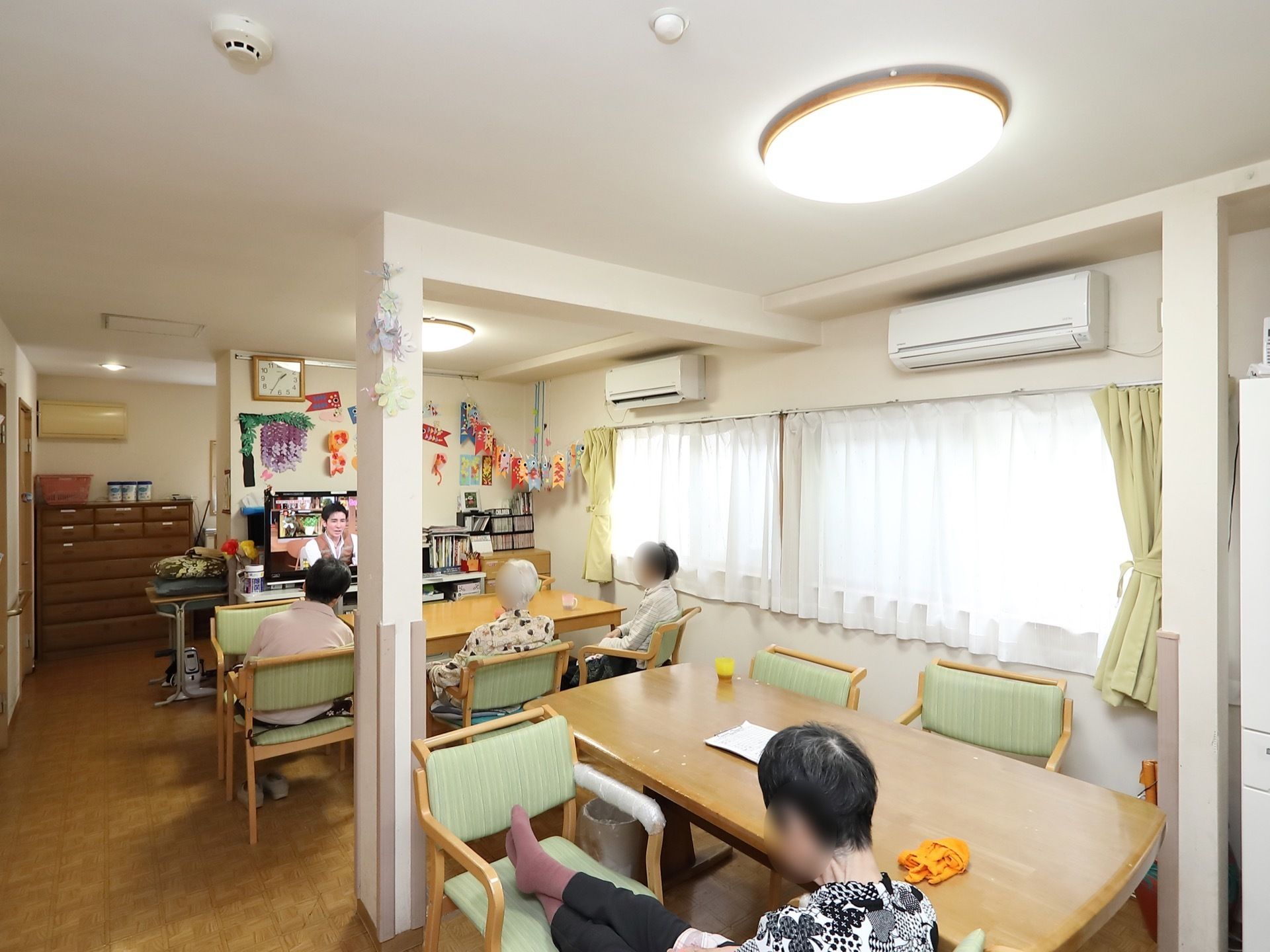 食堂にはテーブルと椅子が用意されており、入居者様がテレビを鑑賞しながらそれぞれ過ごしている。壁には季節に応じた制作物がレイアウトされている。
