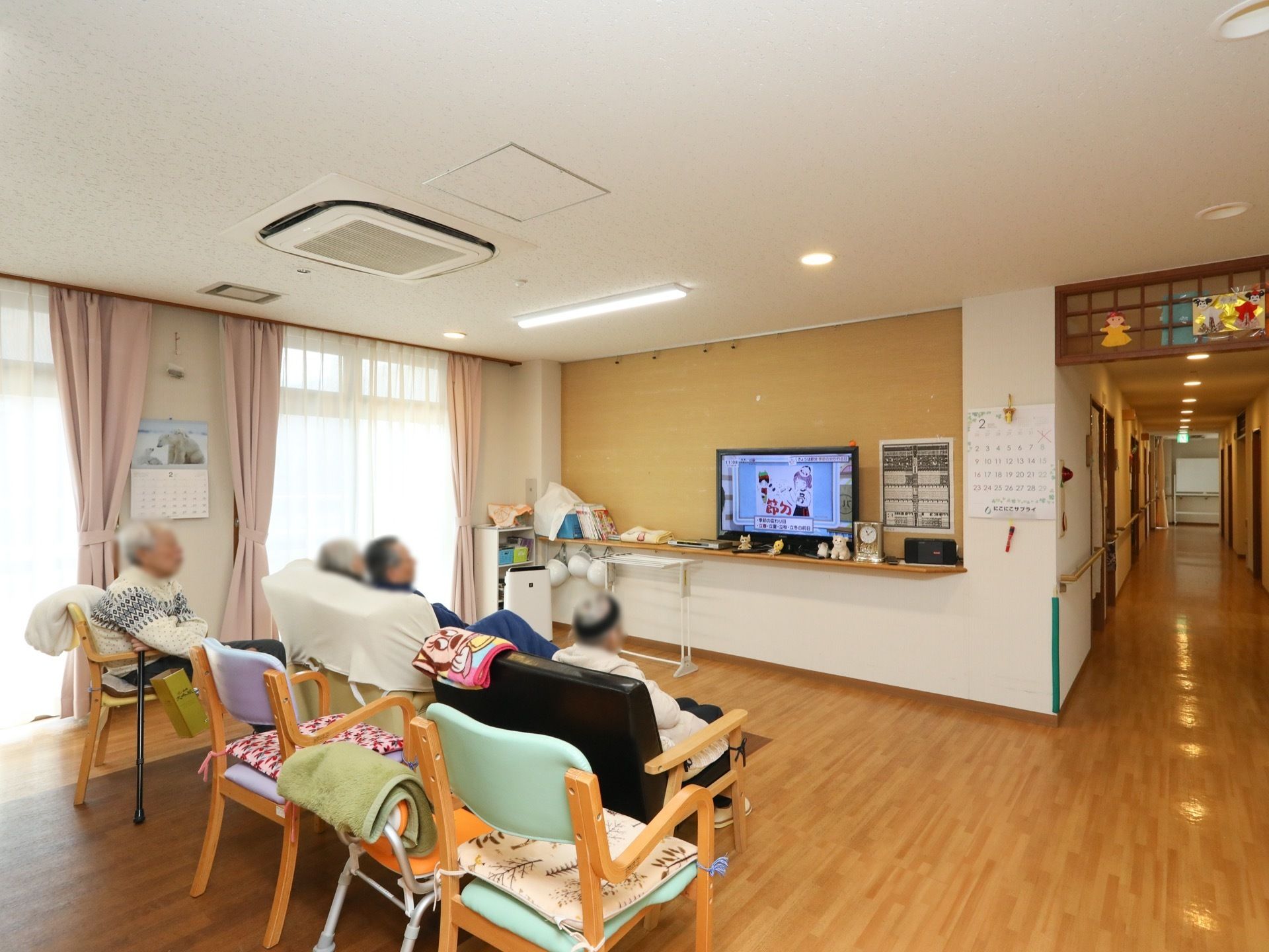 自然光が差し込む共有スペースは、通路から仕切りなく繋がった開放的な空間である。テレビや空気清浄機が置かれ、その前に椅子が並べられている。