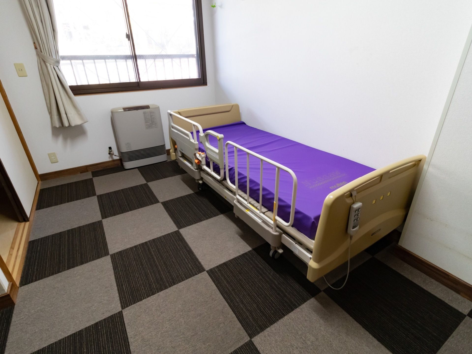 ワンルームタイプの部屋になっている。介護用ベッドが置かれており、窓にはカーテンが掛けられている。床にはカーペットが敷かれている。