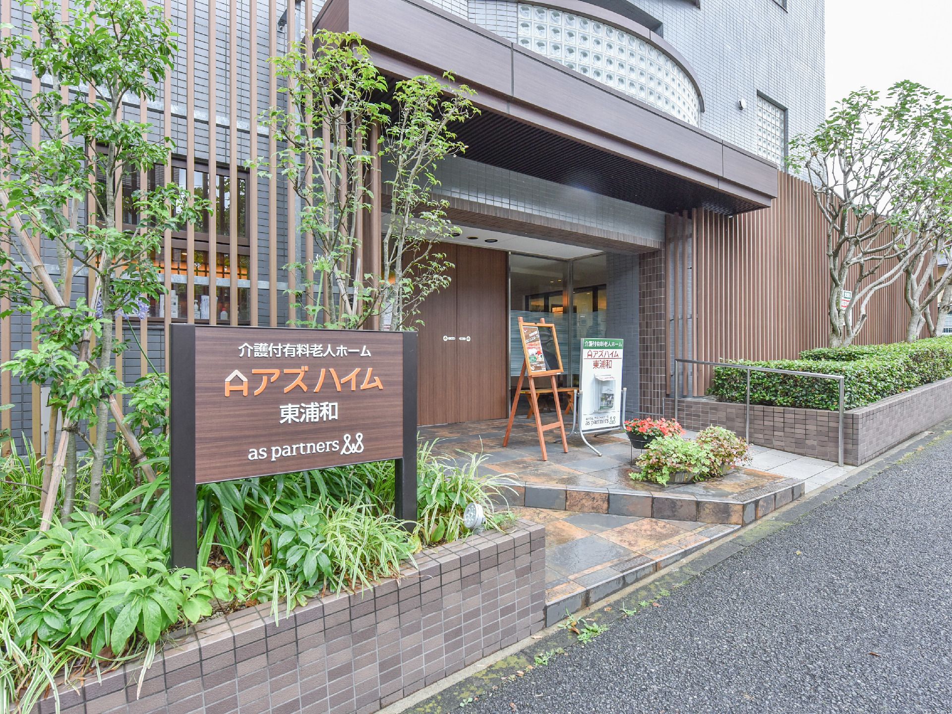 玄関入り口には植え込みがあり、草木が生えている。植え込みには施設名や運営会社名が書かれた看板がある。