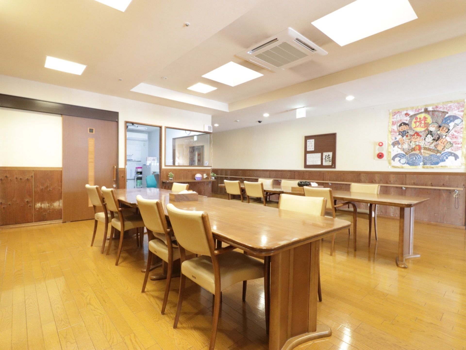 食堂では４人掛けのテーブルと椅子が２列に並べられている。壁には七福神の絵や書の作品が掛けられている。
