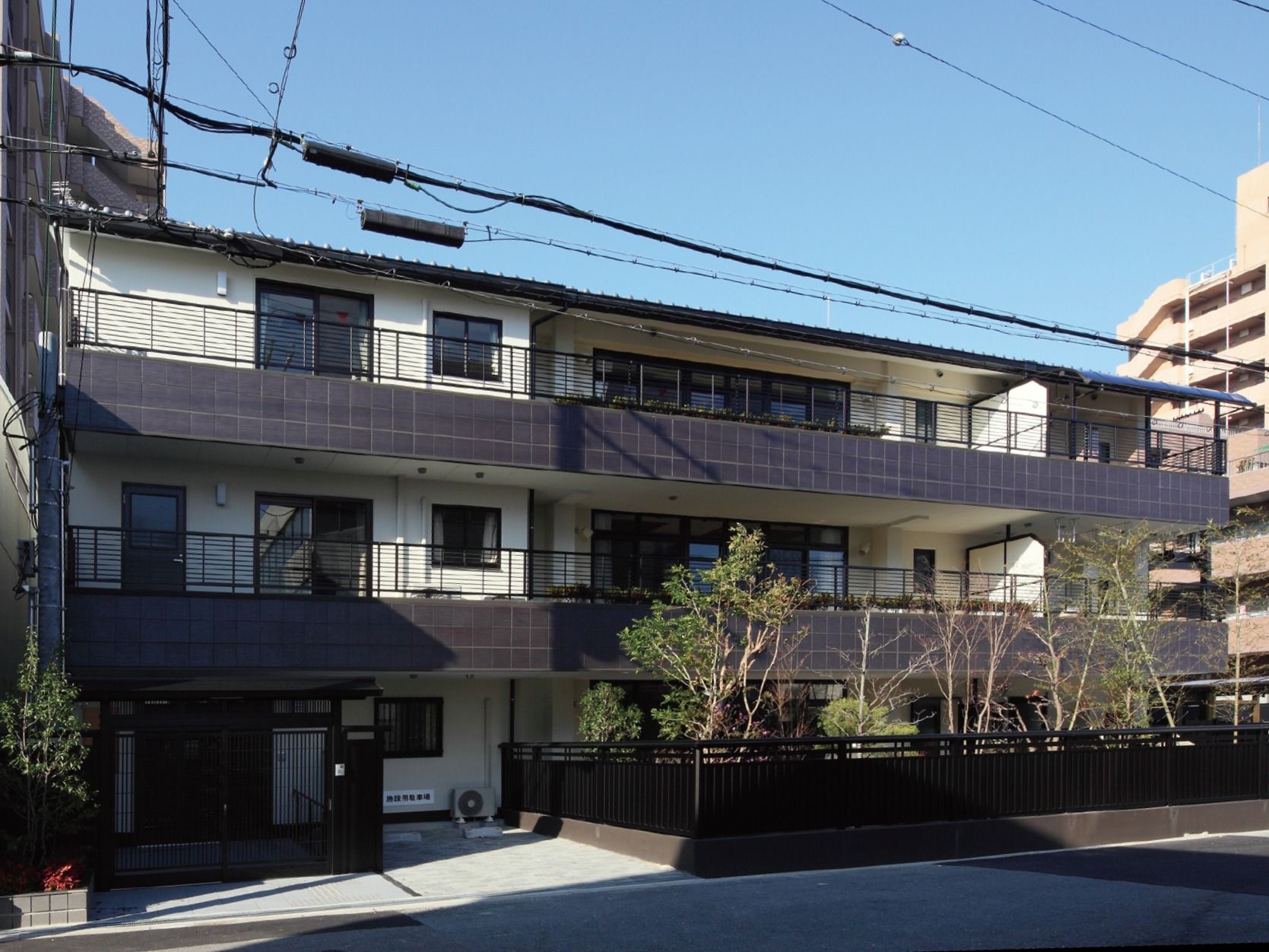 道路に面した3階建ての施設が建っている。外壁は白色であり、ベランダ部分は茶褐色のタイル張りのようになっている。