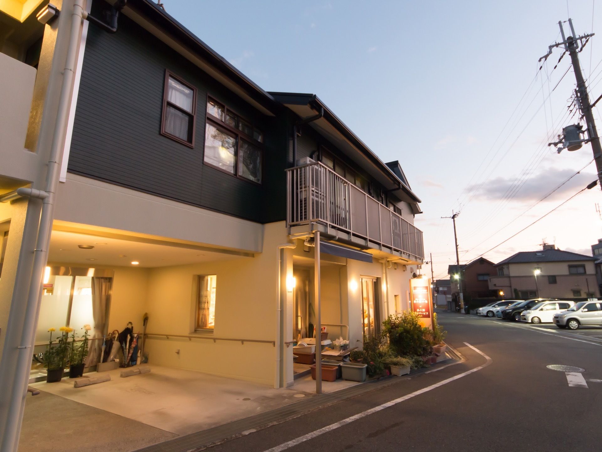 周囲に高い建物がない、夕暮れ時の2階建て施設の様子である。駐車場を完備し、照明器具には明かりが点けられている。