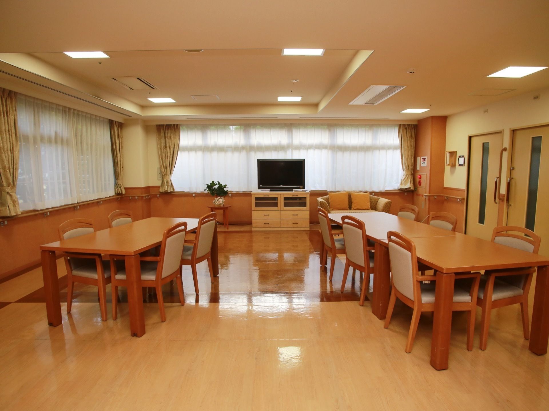 広間に、複数のダイニングセットが並べられている。窓側に大型の液晶テレビや二人掛けソファが置かれている。