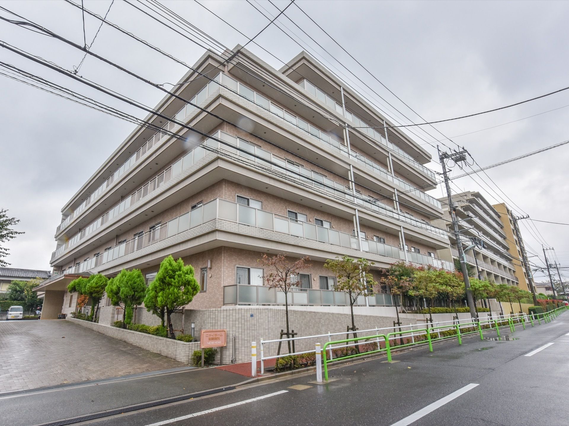 ベージュ系の色でまとめられた落ち着いたデザインの外観である。建物の左横には広いスペースがあり、壁際に緑の木々が植えてある。