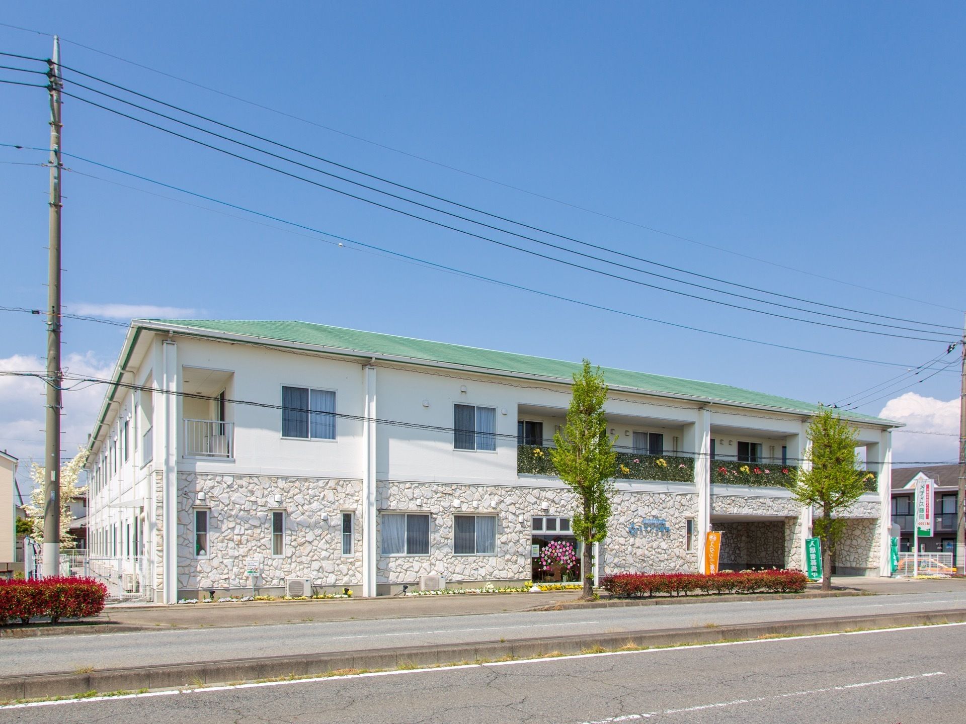 一階部分が石壁調で、緑の屋根の２階建てである。２本の大きな木と赤い低木が、道路沿いに植えられている。