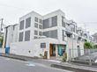 グレーと白の3階建ての建物。玄関は道路沿いではない。スタッフが手書きした看板を設置。他には、鉢植えもある。