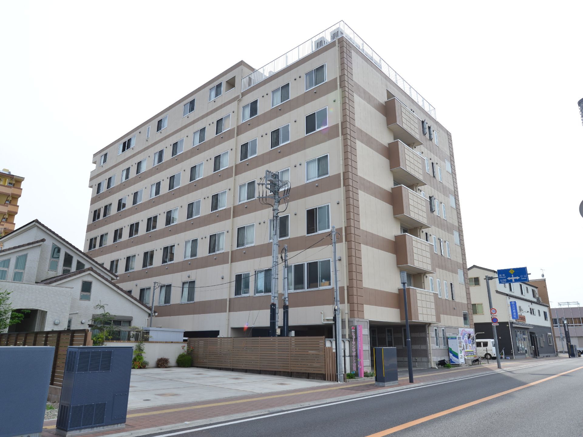 道路に面した、茶色とベージュのツートンカラーの建物。7階建てで、一部屋上になっている。奥行き方向が長い。
