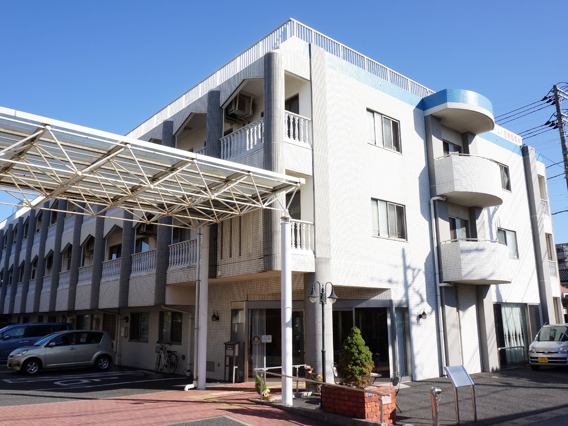 白い外観の施設にあるベランダの柵は洋風でおしゃれである。駐車場から入り口までは屋根付き通路があり、雨の日も安心である。