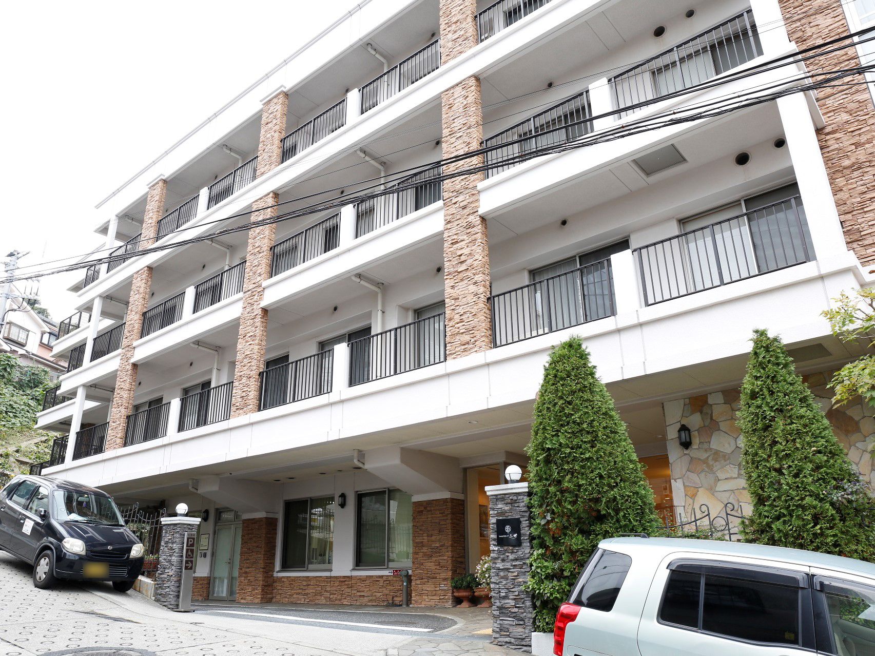 白と茶色でまとめられた4階建ての外観である。手前の道路は少し坂道になっていて、道路わきに2台の車が置いてある。