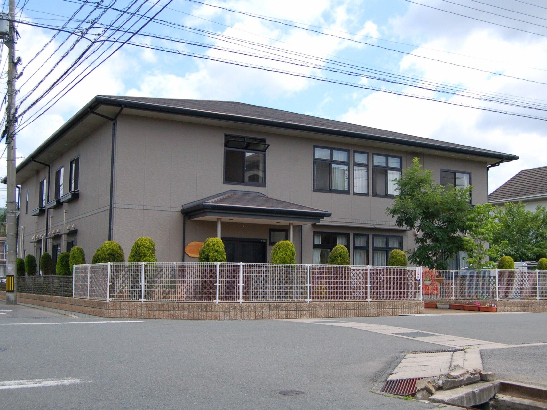 グレー系の外観の２階建ての建物は日当たりがいい場所に建っていて、敷地の周りにはフェンスがある。