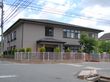 グレー系の外観の２階建ての建物は日当たりがいい場所に建っていて、敷地の周りにはフェンスがある。