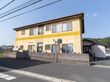 道路沿いにブロック塀に囲まれた、黄色の壁の２階建ての建物がある。建物の周囲には山や住宅、田んぼなどの景色が広がっている。