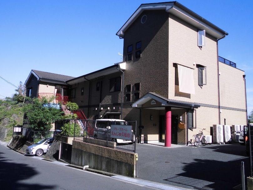 ブラウン系の建物は3階建てで、周りには草木が植えられ、日当たりのいい場所に建っている。