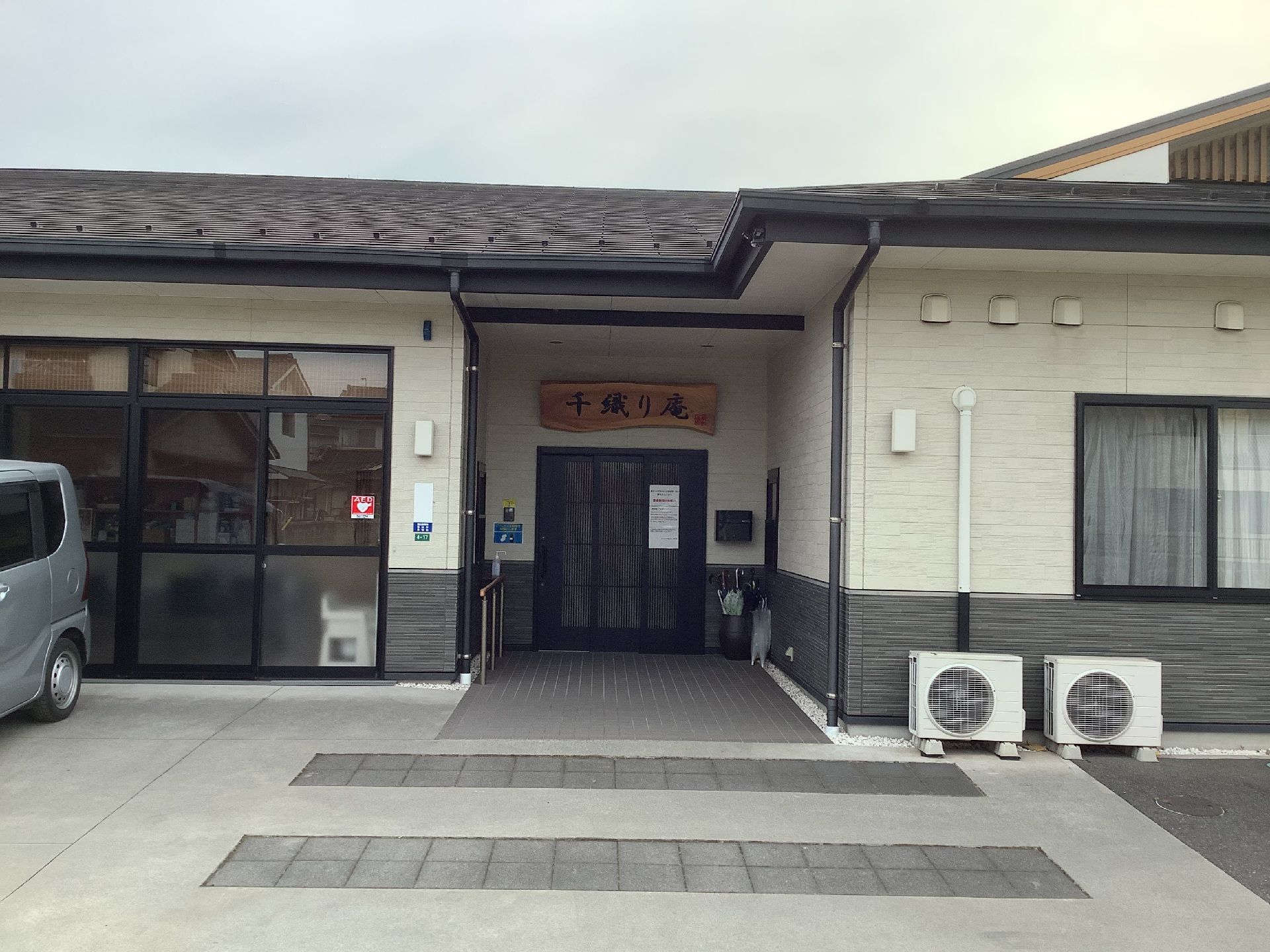 白い外壁の平屋建ての建物の前には車を止める駐車場があり、玄関にはホーム名が書かれている木の板が貼られている。