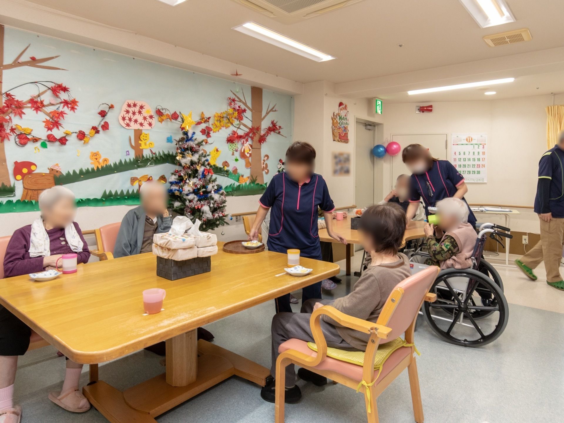 おやつタイムの様子。リビングで入居者様とスタッフが会話を楽しんでいる。壁一面には季節感のある装飾が施されている。