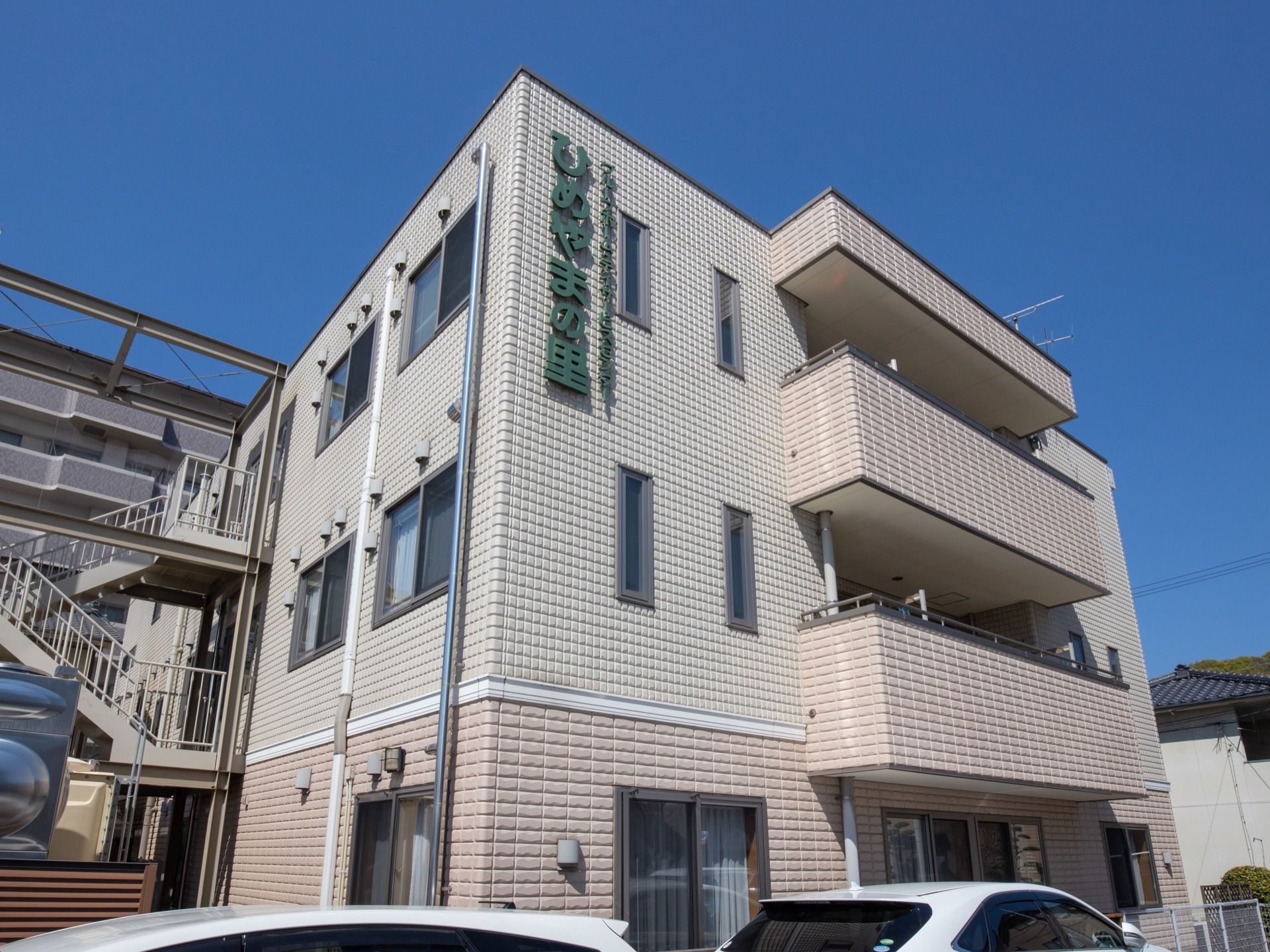３階建ての施設には、外階段とベランダが設置されている。施設名が大きく外壁に記されている。建物裏には、駐車スペースが設けられている。