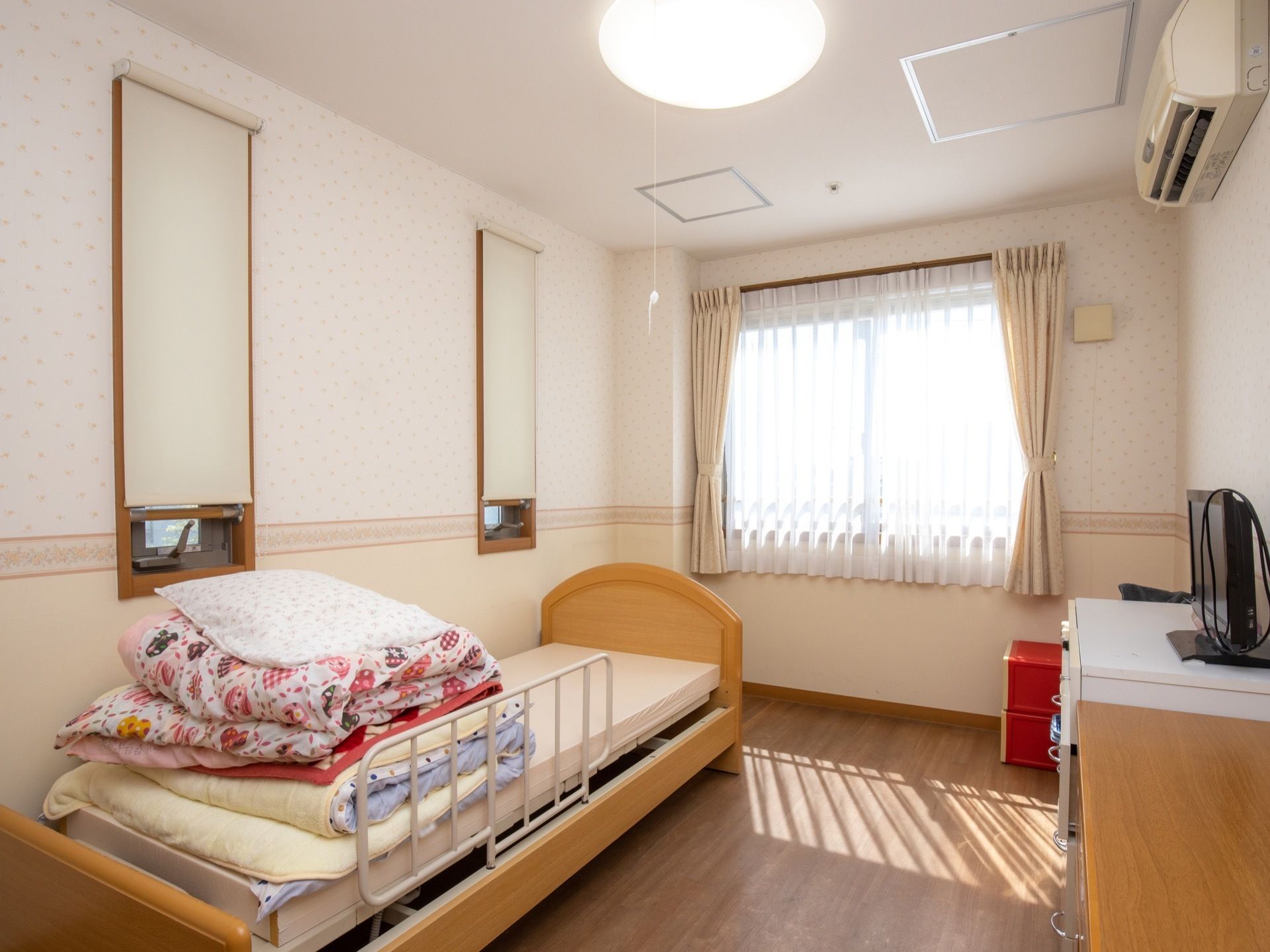 外光が差しこむ窓が設けられた居室である。エアコンやベッドが設置されており、収納家具が壁際に置かれている。