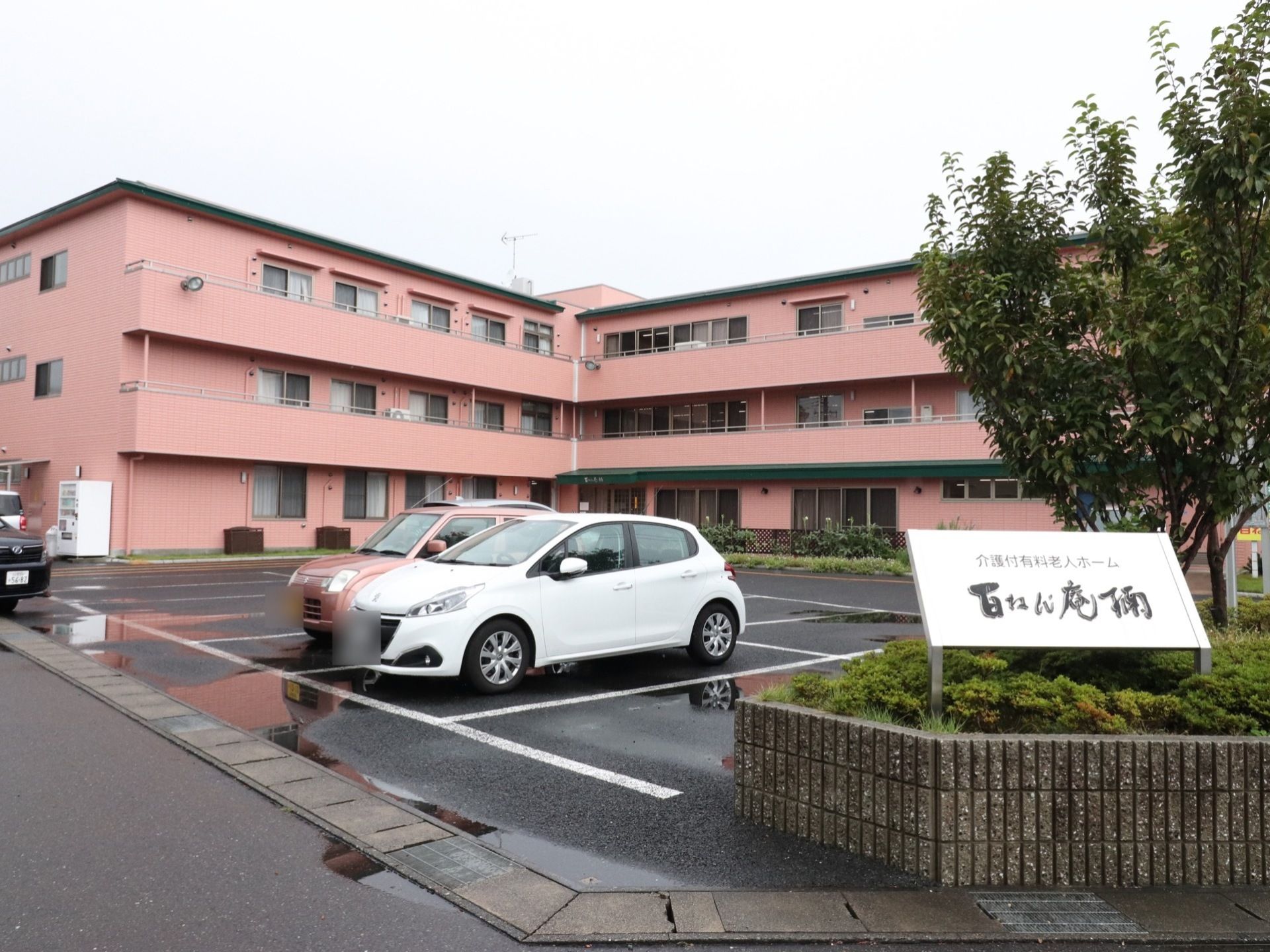 3階建てとなっている施設外観の手前には駐車場があり、何台かの車が止まっている。入り口には看板がある。
