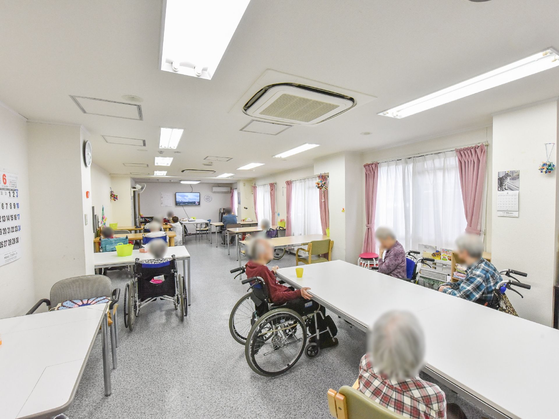 長テーブルが複数並べられている。周りには入居者が椅子や車いすに座っている。大きなテレビが置かれている。