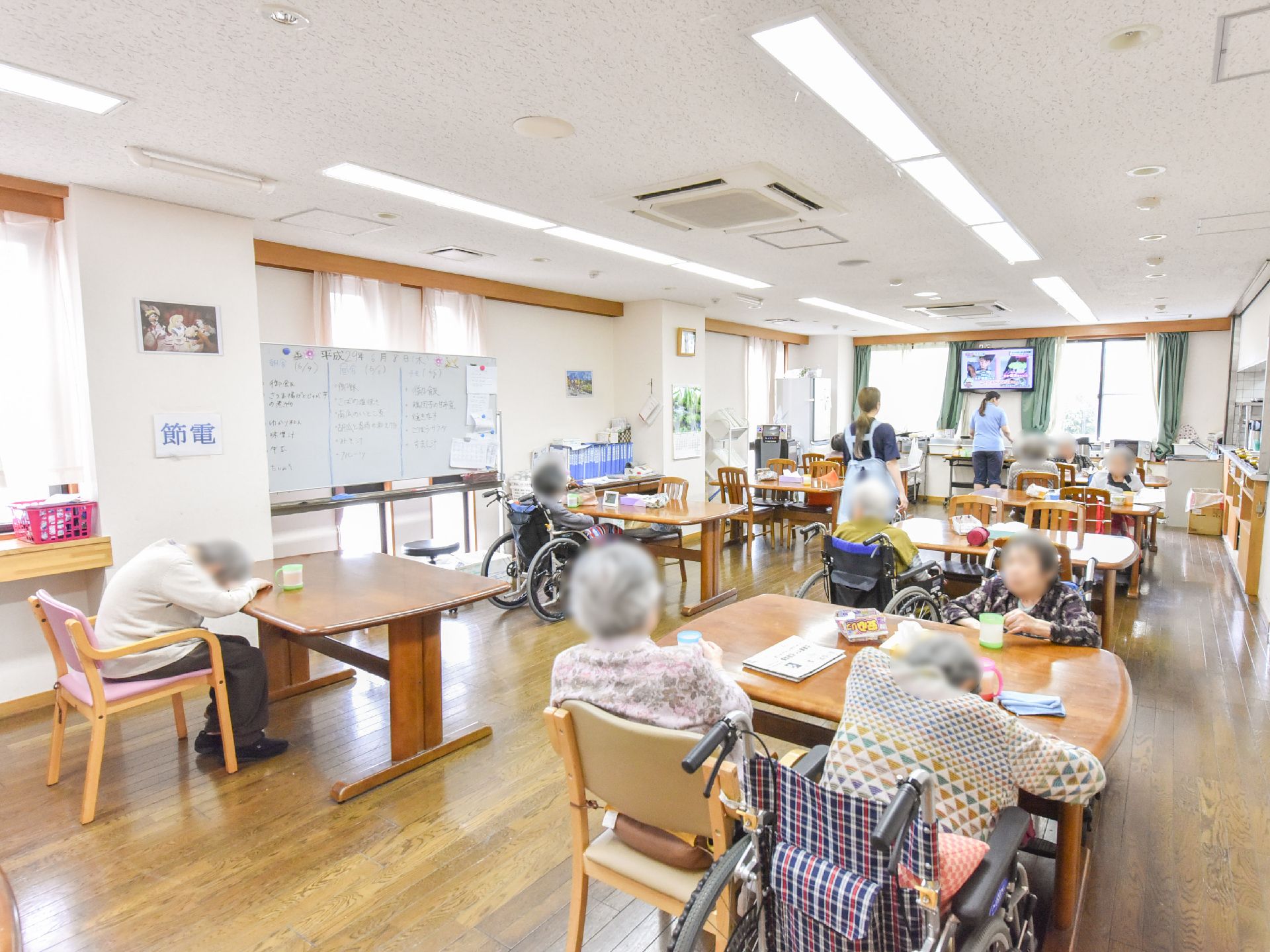 テーブルと椅子が並べられた部屋には入居者たちが座っている。テーブルにはコップが置かれている。前方にはテレビもある。