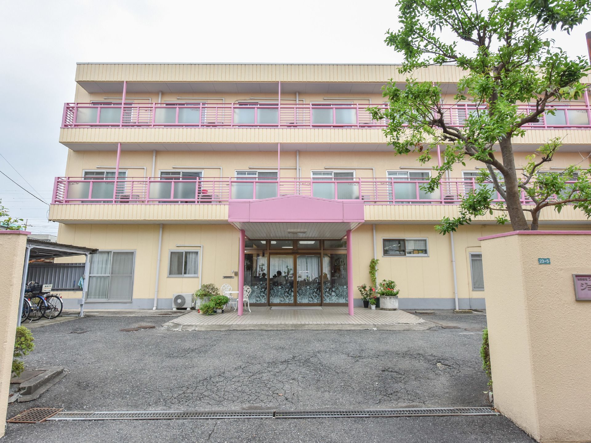 3階建ての建物で、ベージュとピンクの外観になっている。敷地内は広々としていて、駐輪場があり木も植えられている。