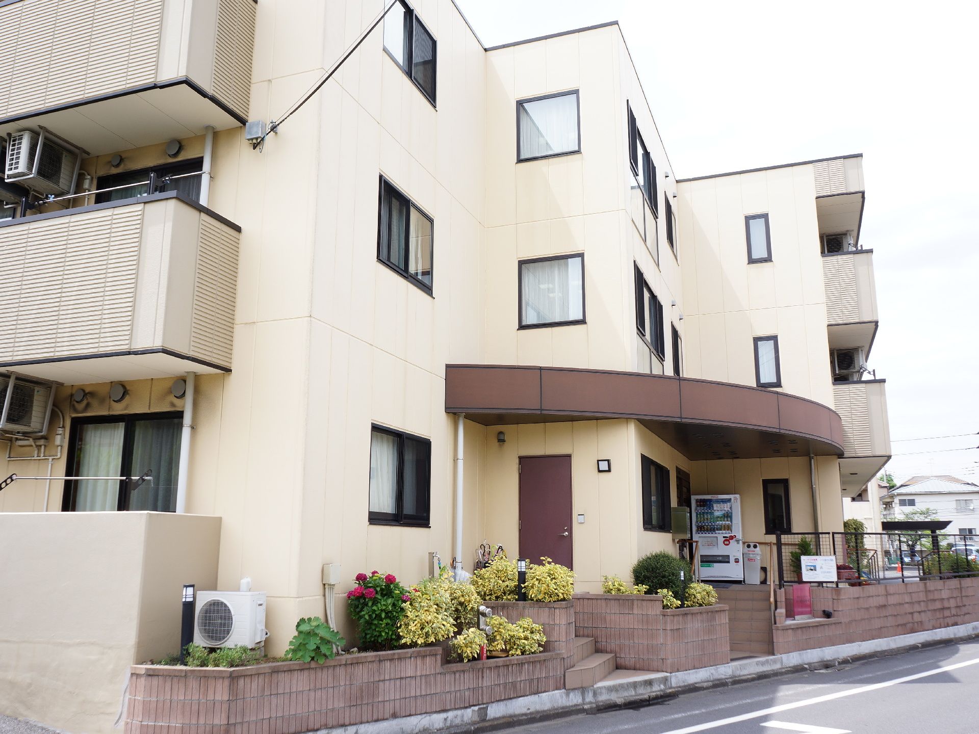 クリーム色の外壁の3階建ての建物になっている。道に面しており茶色の生垣がたち、玄関近くに自動販売機もある。